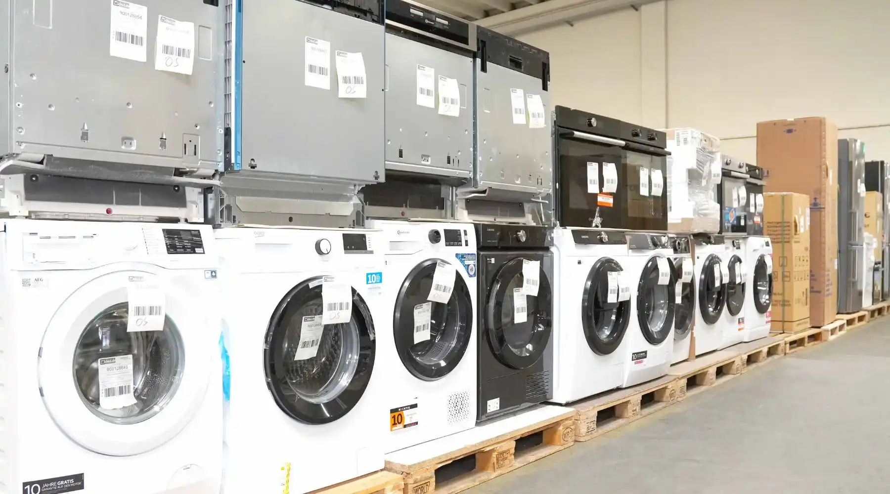 Verschiedene Elektrogeräte wie Waschmaschinen, Trockner und Einbaugeräte auf Paletten im Lager – ideal für alle, die Elektrogeräte kaufen möchten.