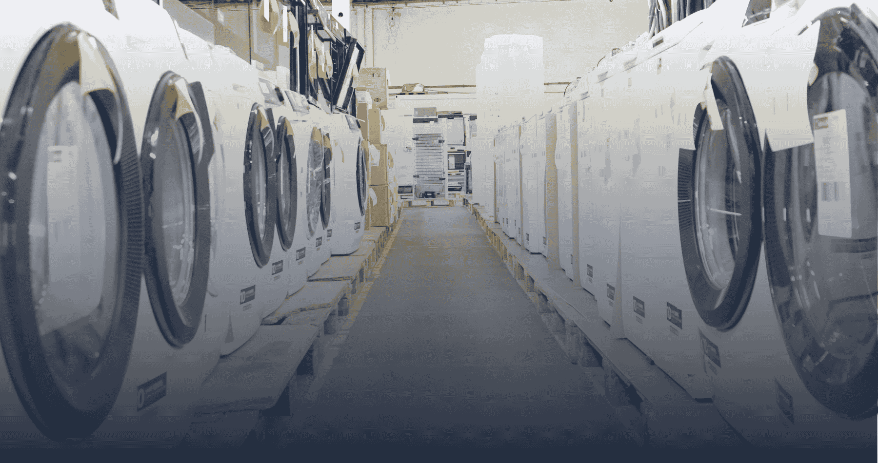 Reihe von auf Paletten stehenden Waschmaschinen, die in einer Lagerhalle eng gegenüber angeordnet sind. Die Geräte stammen aus Retourenbeständen und tragen Etiketten zur Identifizierung. Der mittige Gang führt in Richtung weiterer gestapelter Haushaltsgeräte. Das Bild vermittelt den professionellen Umgang mit Retourenware, wie ihn Händler bei elvinci erwarten können.